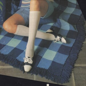 May include: A blue and white checkered crochet throw rug with fringe. The rug is on a tiled floor and a person is sitting on it wearing white knee-high socks and black and white shoes.