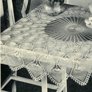 May include: A white crocheted tablecloth with a circular design in the center and a scalloped edge. The tablecloth is laid on a white table with a vase of flowers in the background.