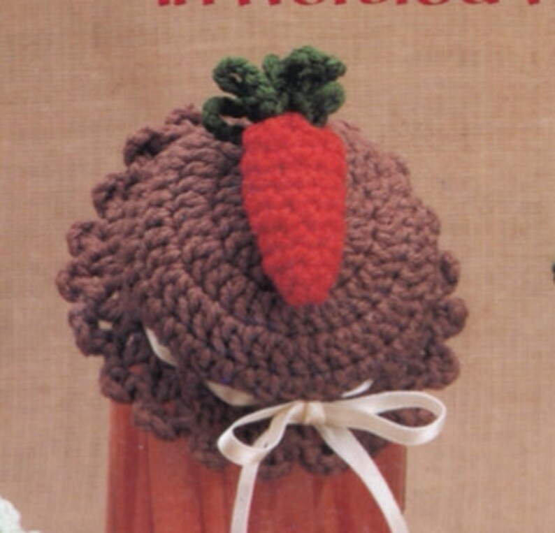 May include: A brown crocheted jar lid with a crocheted carrot and green leaves on top. The lid is tied with a white ribbon.