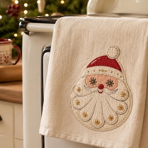May include: A cream-colored kitchen towel featuring an embroidered Santa Claus face. The Santa design includes a red hat with a white pom-pom, a pink face, a white mustache, and a white beard accented with gold-colored star-shaped stitching details.