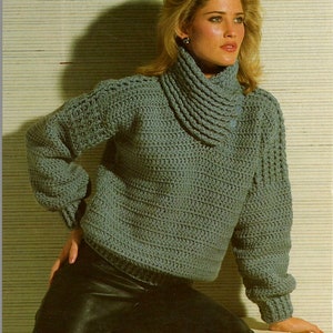 May include: A gray crocheted sweater with a turtleneck and a button detail. The sweater is shown on a model wearing black pants.
