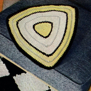May include: A handmade, triangular crochet doily with a yellow, white, and black color scheme. The doily is placed on a blue upholstered chair. The doily's design features concentric triangles, with a black border and a yellow center.