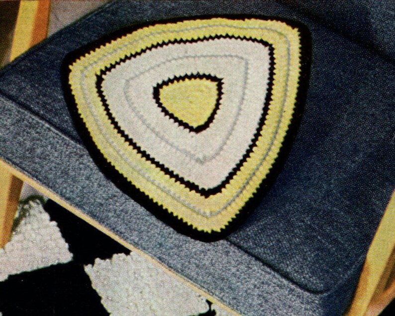 May include: A handmade, triangular crochet doily with a yellow, white, and black color scheme. The doily is placed on a blue upholstered chair. The doily's design features concentric triangles, with a black border and a yellow center.