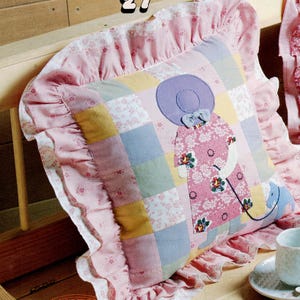 Puede incluir: Un cojín decorativo de patchwork con un borde rosa con volantes. El cojín presenta un diseño de patchwork con cuadrados de varios colores pastel. Una aplicación central representa a una mujer con un vestido y sombrero florales, sosteniendo un parasol y un perro azul pequeño.