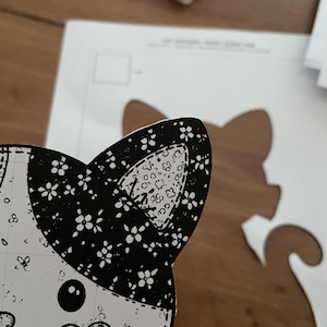 May include: A black and white paper cutout of a cat head featuring a floral pattern on the ears and a heart-shaped nose. The cutout is placed over a white paper template showing the full silhouette of a cat with a curved tail.