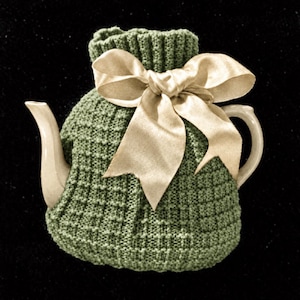 Peut inclure: Une théière en céramique recouverte d'une housse tricotée vert sauge avec un motif à grille texturé. La housse présente un col côtelé et est nouée avec un grand nœud en ruban de satin couleur champagne brillant sur le devant de la théière.