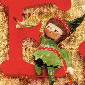 May include: A red wooden letter E with a small elf doll holding a paintbrush and a bucket of paint. The elf is wearing a green and white outfit with red polka dots.