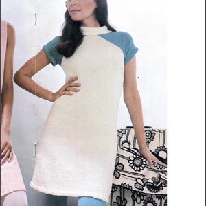 May include: A woman wearing a white dress with blue and green accents. The dress has a high neckline and short sleeves. She is holding a black and white floral patterned suitcase.
