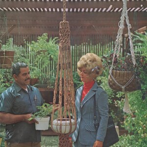 May include: A vintage image featuring a macrame plant hanger with a decorative pot. The brown macrame design is suspended from a wooden structure. A second hanging basket with a woven design is also visible. The scene is set outdoors with lush greenery.