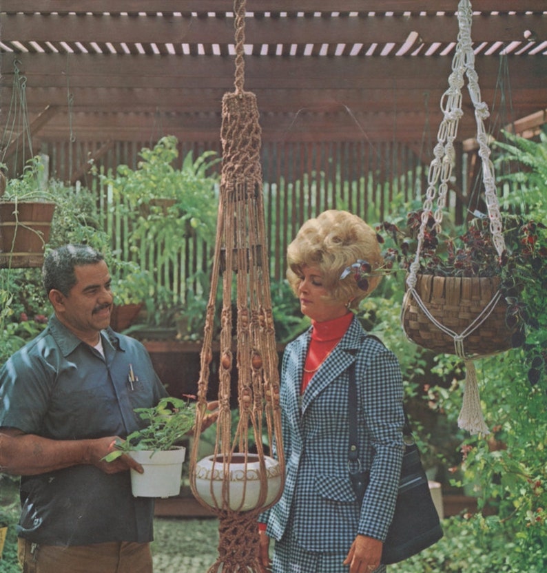 May include: A vintage image featuring a macrame plant hanger with a decorative pot. The brown macrame design is suspended from a wooden structure. A second hanging basket with a woven design is also visible. The scene is set outdoors with lush greenery.