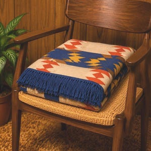 May include: A folded woven blanket featuring a geometric pattern in shades of blue, red, yellow, and cream. The blanket has a textured surface and is finished with a fringe of blue tassels along one edge, displaying a repeating diamond motif.
