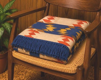 May include: A folded woven blanket featuring a geometric pattern in shades of blue, red, yellow, and cream. The blanket has a textured surface and is finished with a fringe of blue tassels along one edge, displaying a repeating diamond motif.