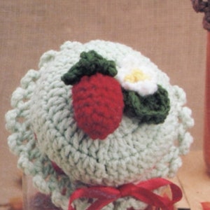 May include: A crocheted jar lid with a strawberry and flower design. The lid is green, white, and red.