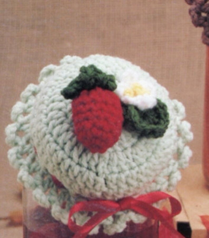 May include: A crocheted jar lid with a strawberry and flower design. The lid is green, white, and red.