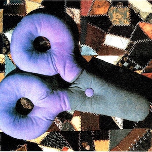 May include: A unique, handmade pillow with two purple donut-shaped cushions connected to a gray rectangular base. The pillow is set against a patchwork quilt background with various fabric patterns and colors.