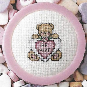 Puede incluir: Un aro de bordado de punto de cruz rosa y blanco con un osito de peluche que sostiene un corazón que dice "Be Mine". El aro está rodeado de corazones de caramelo rosa y blanco.