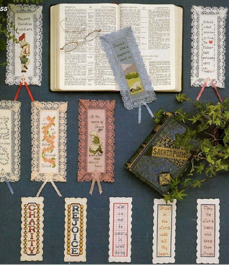 May include: A collection of 10 handmade bookmarks with lace trim and cross-stitch designs. The bookmarks feature religious quotes and images, including a shepherd, a girl, and flowers. The bookmarks are displayed on a blue background with a book and greenery.