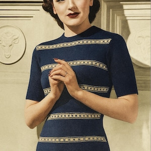Op de afbeelding: Een marineblauw gebreid topje met korte mouwen en horizontale strepen met een herhalend goudkleurig patroon. De top heeft een hoge halslijn. De vrouw op de foto draagt een ring aan haar vinger.