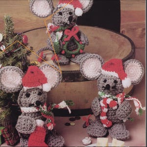 May include: Three crocheted mouse ornaments wearing Santa hats. The mouse on the left is holding a candy cane, the middle mouse is holding a wreath, and the mouse on the right is holding a candy cane.