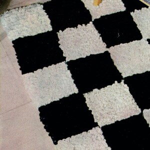 May include: A black and white checkerboard rug with a textured, looped pile. The rug is rectangular and features alternating black and white squares. The rug is placed on a light-colored wooden floor, with the legs of a chair visible.