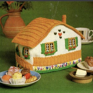 May include: A house-shaped tea cozy featuring a white knitted body with orange roof and base trim. The design includes green shutters, small windows with red flower boxes, and a floral border at the bottom. A small chimney sits on the roof.