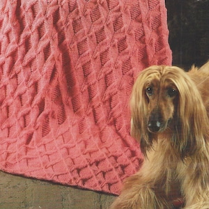 May include: A pink knitted throw with a cable knit pattern. A long-haired dog is sitting in front of the throw.