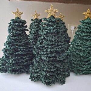 May include: A collection of four cone-shaped Christmas trees crafted from dark green textured yarn, each topped with a small, shimmering gold star. The trees feature a layered, ruffled appearance created by the crochet stitch pattern.