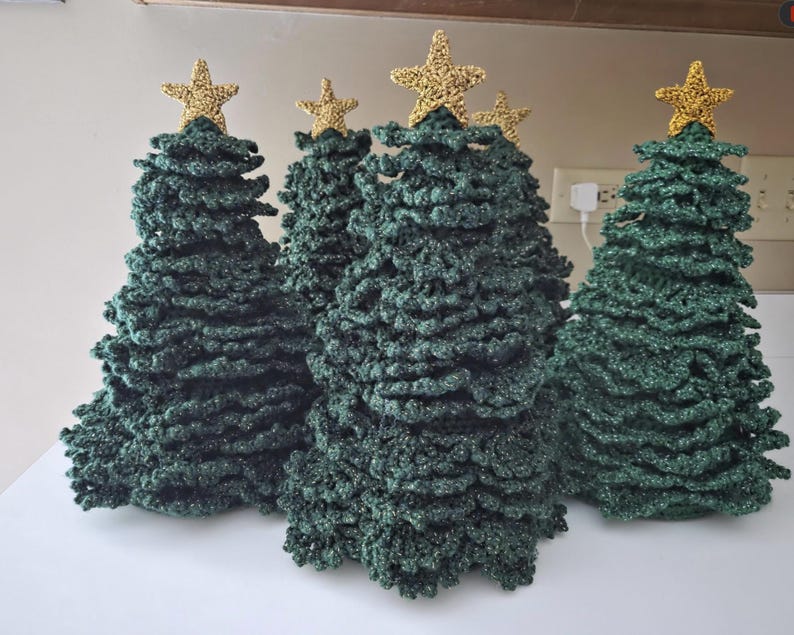 May include: A collection of four cone-shaped Christmas trees crafted from dark green textured yarn, each topped with a small, shimmering gold star. The trees feature a layered, ruffled appearance created by the crochet stitch pattern.