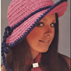 May include: A pink crocheted hat with a dark blue trim and a matching braided tie. The hat has a wide brim and is worn by a person with long brown hair. The hat is a vintage style.