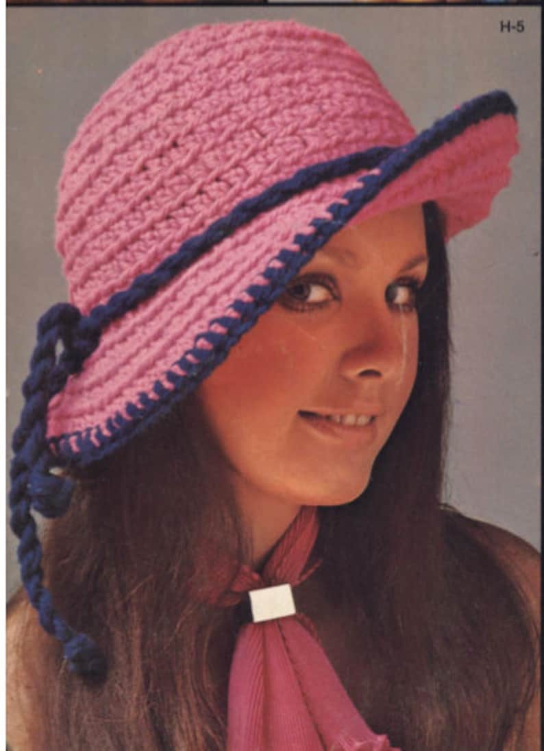 Pu&ograve; includere: Un cappello rosa all'uncinetto con finiture blu scuro e un cordino intrecciato abbinato. Il cappello ha un'ampia tesa ed &egrave; indossato da una persona con lunghi capelli castani. Il cappello &egrave; in stile vintage.