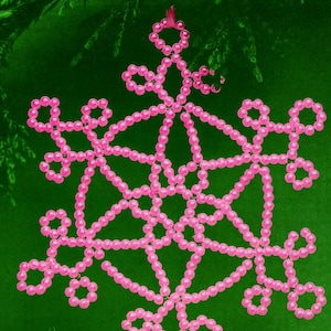 Könnte beinhalten: Ein rosa Perlen-Schneeflocken-Ornament vor einem grünen Hintergrund. Die Schneeflocke besteht aus kleinen, runden Perlen, die zu einem symmetrischen Design aufgefädelt sind. Das Ornament hat eine Schlaufe zum Aufhängen.