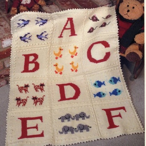 Könnte beinhalten: Eine weiße gehäkelte Babydecke mit einem bunten Alphabet-Design. Jeder Buchstabe ist von einem anderen Tier oder Objekt umgeben, darunter Vögel, Enten, Mäuse, Katzen, Fische und Elefanten.