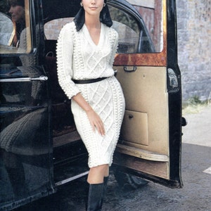 May include: A woman wearing a white cable knit dress with a black belt and black boots. She is standing next to a black car with the door open. She is wearing a black headscarf.