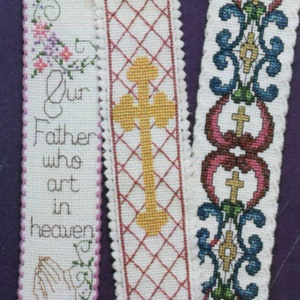 Peut inclure: Trois marque-pages en tissu blanc avec des motifs de point de croix. Le premier marque-page a le texte "Notre P&egrave;re qui es aux cieux" et une paire de mains en pri&egrave;re. Le deuxi&egrave;me marque-page a une croix jaune sur un motif de grille rouge et blanche. Le troisi&egrave;me marque-page a un motif floral bleu et vert avec un c&oelig;ur rouge et une croix.