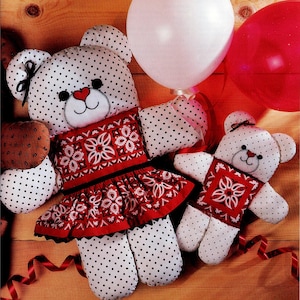 Può includere: Due orsacchiotti fatti a mano con tessuto bianco e pois neri. Gli orsi indossano abiti a fantasia rossi e bianchi. Un palloncino bianco e un palloncino rosso sono sullo sfondo. Un nastro rosso è anche nell'immagine.