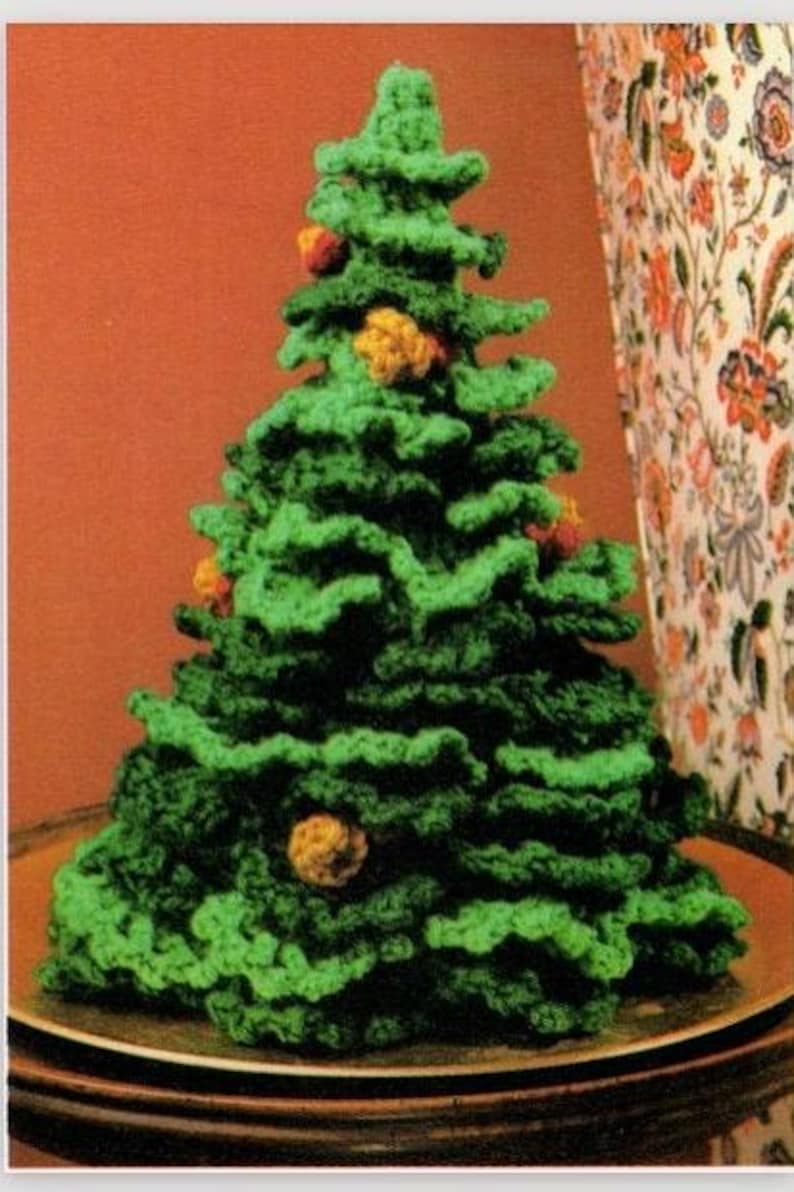 May include: A conical crochet Christmas tree crafted from green yarn with ruffled, layered branches. The tree is decorated with small, round, textured ornaments in shades of yellow and orange scattered throughout the foliage.