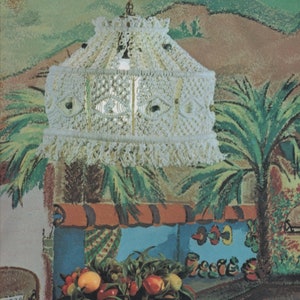 May include: Cream-colored macrame pendant light with a fringed bottom. The lamp hangs from a brass-colored chain. The background features a painted mural of a tropical scene with palm trees and a fruit display.