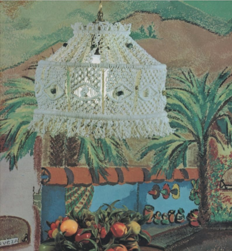 May include: Cream-colored macrame pendant light with a fringed bottom. The lamp hangs from a brass-colored chain. The background features a painted mural of a tropical scene with palm trees and a fruit display.