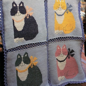 Könnte beinhalten: Eine gehäkelte Decke mit vier Quadraten, die verschiedene farbige Katzen zeigen. Jede Katze trägt eine Schleife und ein weißes Halsband. Die Decke ist hellblau mit einem violetten Häkelrand.