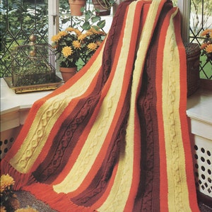 May include: A striped knit afghan with a cable knit pattern in shades of orange, yellow, and brown. The afghan has fringe on the bottom edge. The afghan is draped over a chair in a sunroom.