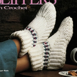 May include: A pair of white crochet slipper boots featuring a ribbed texture and a decorative band with small red and green accents. The text SLIPPERS In Crochet and Slipper Boots is visible at the top and bottom of the image.