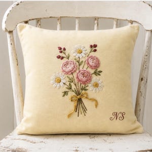 May include: A square, pale yellow throw pillow featuring a central embroidered bouquet of pink roses, white daisies, and green foliage tied with a mustard yellow ribbon. The letters NS are embroidered in brown in the bottom right corner.