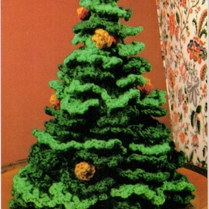 May include: A crocheted Christmas tree in various shades of green yarn, featuring a layered, ruffled texture. Small, round, textured ornaments in shades of yellow, orange, and red are attached to the branches of the conical-shaped tree.