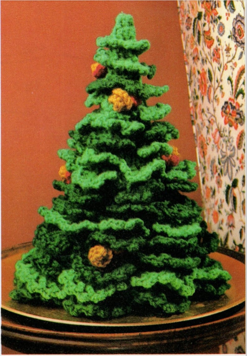 May include: A crocheted Christmas tree in various shades of green yarn, featuring a layered, ruffled texture. Small, round, textured ornaments in shades of yellow, orange, and red are attached to the branches of the conical-shaped tree.