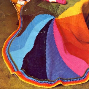 May include: A colourful crocheted blanket with a rainbow pattern. The blanket is made with different shades of blue, green, yellow, orange, pink, and red yarn. The blanket has a scalloped edge.