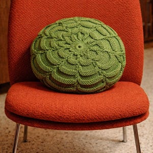 May include: A round, green knitted cushion with a detailed floral pattern sits on a red upholstered chair. The chair has a curved back and chrome legs. The cushion is approximately 35 cm in diameter.