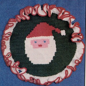 May include: A crocheted Christmas decoration depicting Santa Claus. The design features a green circular base with a white Santa face, a red hat, and a ruffled white and red border. The background is a solid blue.