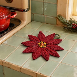 May include: A crocheted poinsettia-shaped potholder featuring deep red petals with textured stitching and a central cluster of small yellow and green knots. The floral-shaped kitchen accessory includes a small green loop for hanging purposes.