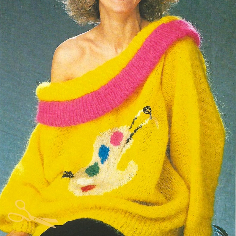 May include: A yellow fuzzy sweater with a pink trim around the neckline and a white paint palette design on the front.