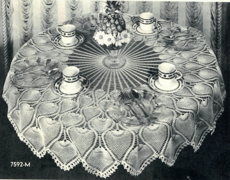 May include: A white crocheted tablecloth with a scalloped edge and a circular design in the center. The tablecloth is set for a tea party with four cups and saucers, a pineapple centerpiece, and silverware.